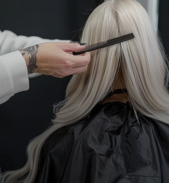 Coiffeur coiffant doucement de longs cheveux blonds platine, vu de dos, préparation avant pose ou coiffage